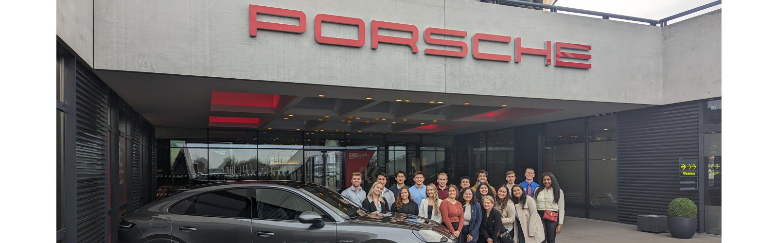 Students are outside of a Porsche building where there is a Porsche car in the foreground. The students are close together and smiling at the camera.